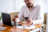 Man sitting at kitchen table looking at credit card and phone with laptop and papers