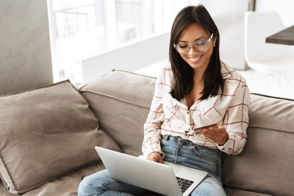A young woman sits on a couch and makes an online purchase using her credit card after learning how to prevent credit card fraud.