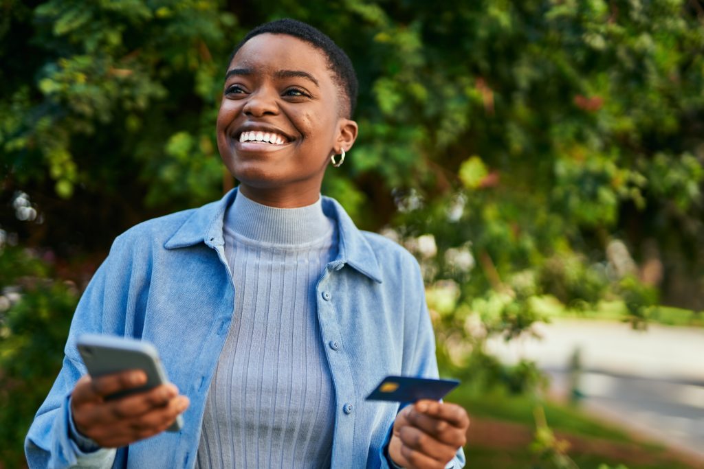 A woman smiles in a park on a sunny day, using her credit card online, enjoying the benefits that come with credit card rewards.