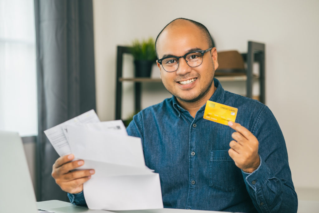 A man sits at a table with papers and a credit card, smiling because he is saving money after a credit card balance transfer.