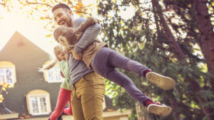 An father is holding a child in their arms, both are mid-air and appear to be playing outside a house surrounded by trees.