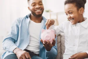 Man holding a piggy bank while his little girl puts a coin into it
