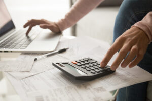 Woman using a calculator and laptop calculating monthly expenses and budgets, sitting at a table with documents.