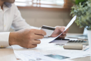 Person holding a credit card and document, with documents and a calculator on a desk.