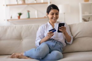 Young woman smiling, sitting on a couch, looking at a smart phone holding a credit debit card.
