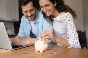 Happy couple sitting at home, putting coins into a piggy bank, using a laptop.