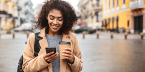 Cheerful young woman wearing coat, walking outside, looking at a smart phone holding a coffee cup.