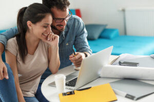 Happy couple sitting at home, looking at laptop with notebooks at desk.