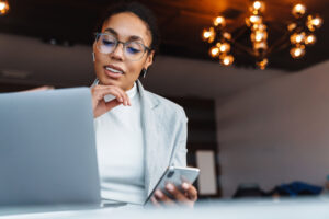 Woman with glasses holding a phone, looking at a laptop.