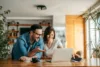 Man leaning over the kitchen table looking at the laptop his wife is looking at. The table has a laptop, coffee cup, laptop, and papers on it.
