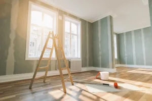 Room under renovation with unpainted walls, ladder, painting tools, and a large window.