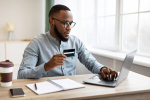 African American man sitting in front of a laptop holding a bank card.