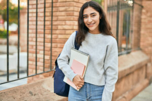 Young girl smiling carrying a backpack holding a notebook.