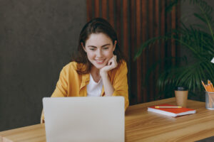 Young woman in a yellow jacket, smiling and sitting at a desk, using a laptop.