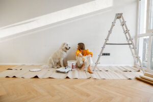 Woman sitting in a empty room with painting supplies, ladder, and a dog.