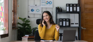 Woman in an office, talking on the phone, sitting at a desk in front of a laptop.