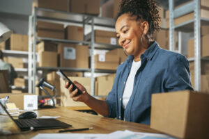 Woman working in a warehouse holding a phone and looking at laptop.