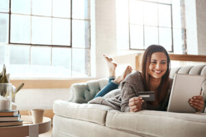 Woman sitting on a couch smiling, looking at electronic device holding a bank card.