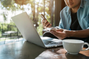 Man sitting in front of a laptop and teacup, holding a bank card and phone.