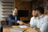 Young couple sitting a desk shaking hands with a bank advisor. The desk has a laptop, contract, and folder on it.