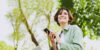 A person holding a smartphone and looking at it, standing outdoors with green trees in the background.
